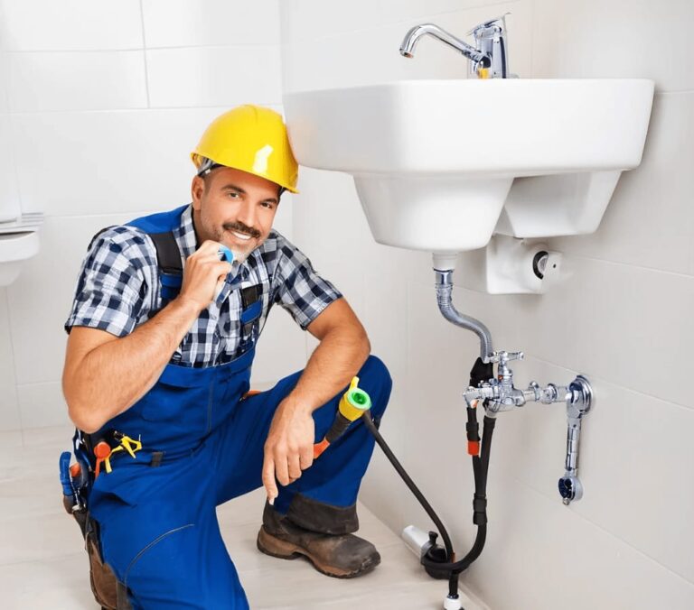 Un plombier patric oliveira portant un casque de sécurité jaune et une salopette bleue travaille sur une installation sous un évier. Il utilise un ruban de plomberie pour sécuriser les tuyaux et accessoires, illustrant un professionnel en pleine intervention.